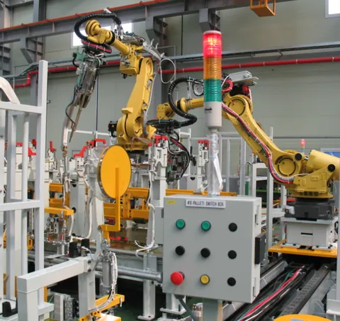 Robotic assembly cell on a factory floor with a control panel and safety fencing